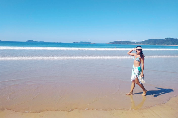 Imagem de uma mulher caminhando descontraída pelas areias da Praia de Geribá de canga e viseira com os chinelos na mão. Praias de Búzios