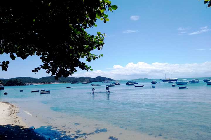 Imagem da Praia da Armação com a escultura dos Três Pescadores ao fundo e vários barcos ancorados.