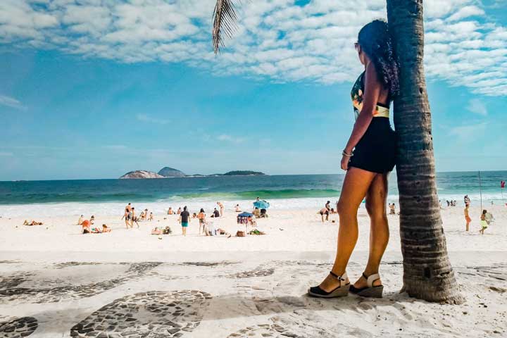 Imagem de uma mulher escorada em um coqueiro admirando a praia de Ipanema