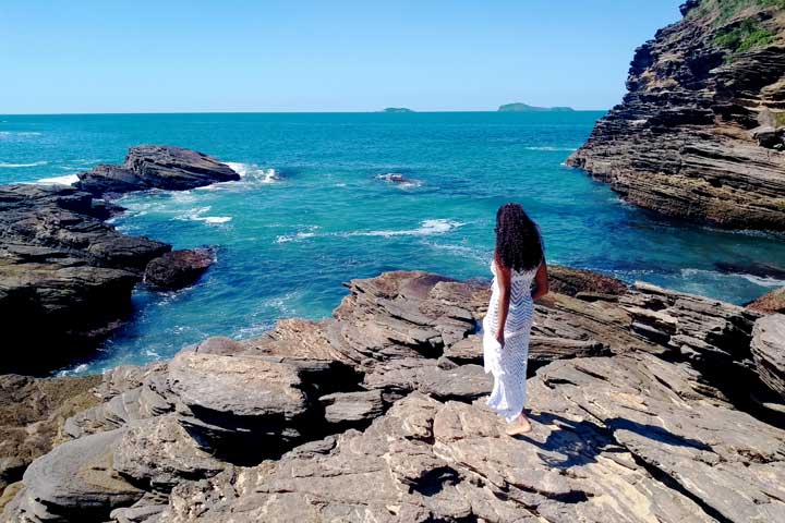 Imagem de uma mulher de de vestido branco caminhando nas rochas da Praia da Ferradurinha em Búzios.