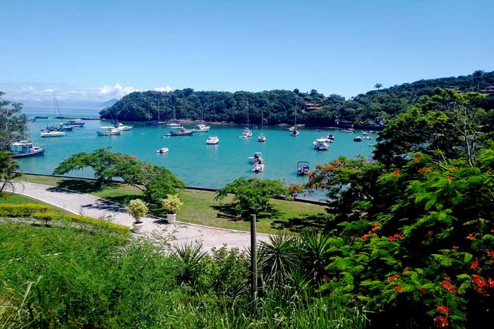 Imagem da Praia dos Ossos vista de um ponto mais elevado, com sua paisagem composta de muito verde que se contrasta com as aguas azuis e os vários barcos ancorados.