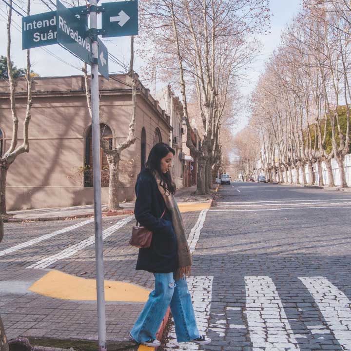 Mulher descontraída e com as mãos no bolso caminhando pelas ruas do Centro Histórico de Colonia del Sacramento, durante um dia frio e com sol.