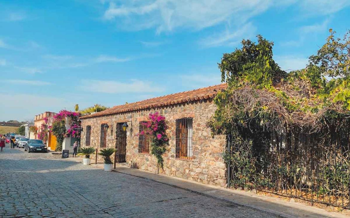 A imagem captura a entrada do Charco Hotel, ladeada por arbustos, em uma charmosa rua pavimentada com pedras.