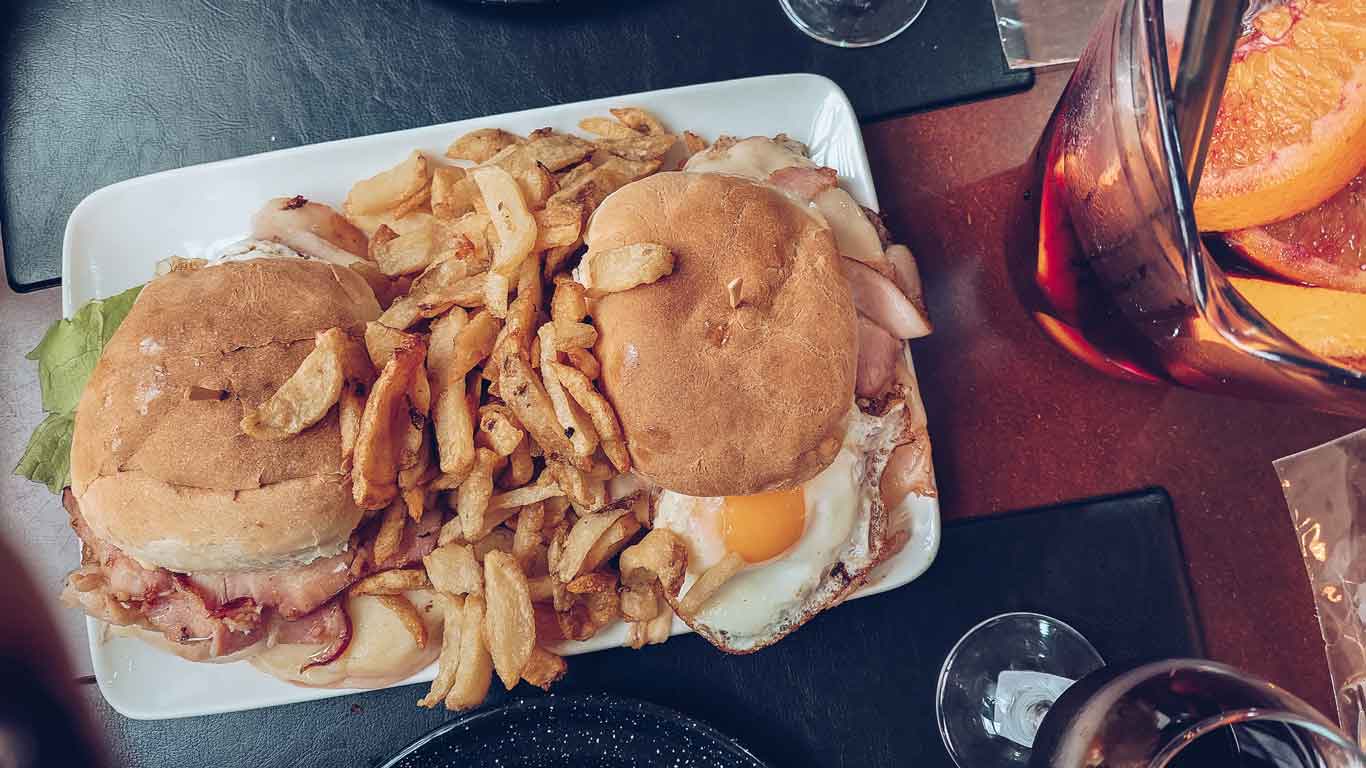 Foto de dois chivitos acompanhados por batatas fritas, ao lado de uma jarra de sangria.