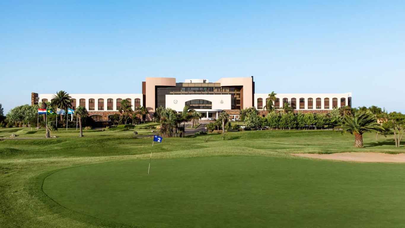 A imponente estrutura do Sheraton Colonia Golf & Spa Resort é retratada, destacando-se pela sua vasta área verde à frente.