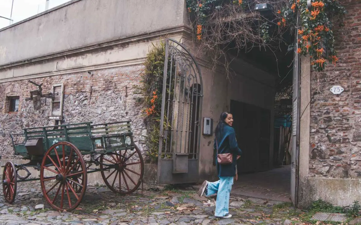 Uma mulher sorridente caminha ao lado de um carro antigo em uma rua de paralelepípedos. O carro está estacionado em frente a uma parede de pedra rústica com uma porta de ferro forjado. Flores laranjas pendem de uma treliça sobre a entrada do edifício.