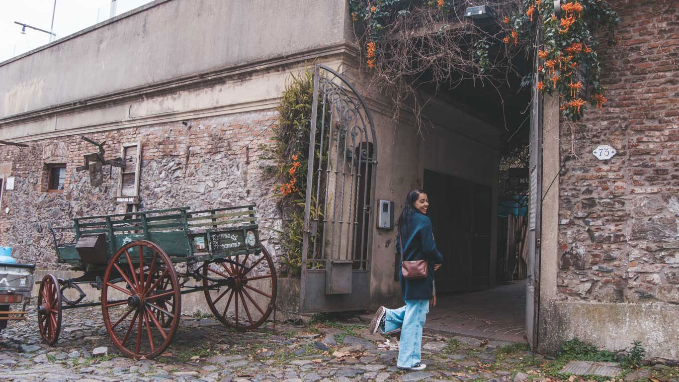 Uma mulher sorridente caminha ao lado de um carro antigo em uma rua de paralelepípedos. O carro está estacionado em frente a uma parede de pedra rústica com uma porta de ferro forjado. Flores laranjas pendem de uma treliça sobre a entrada do edifício.