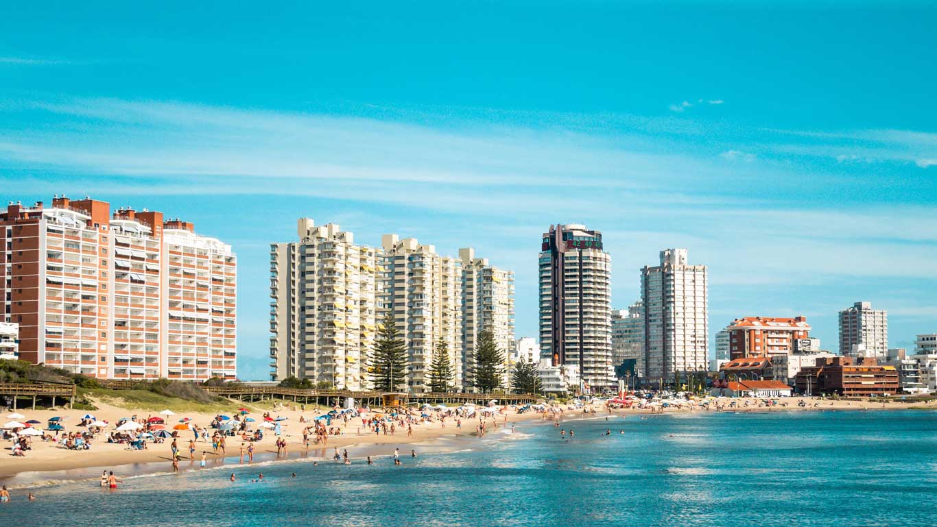 Uma cena da costa de Punta del Este revela um mar de tonalidades azuis, onde pessoas ocupam a extensão das areias.