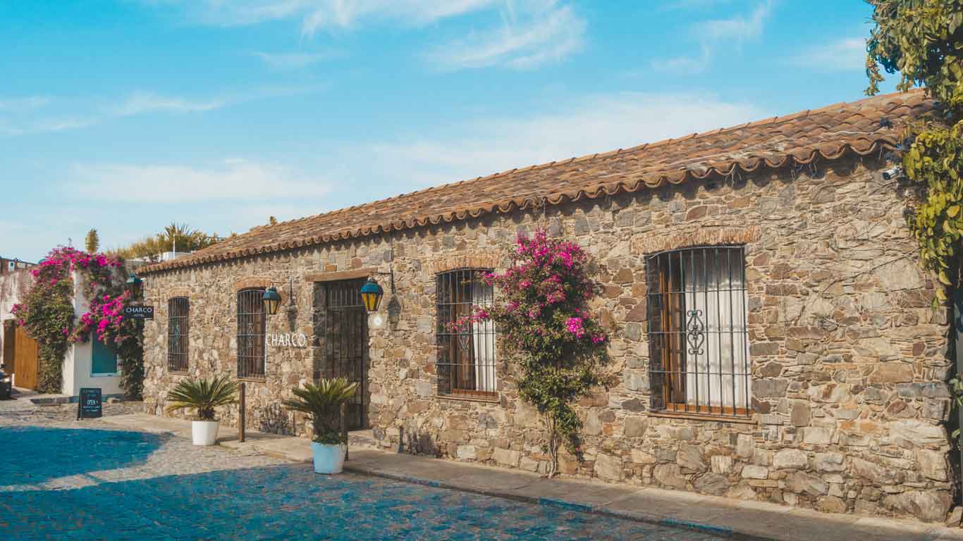 A foto retrata o Charco Hotel, um local aconchegante com sua parede de pedras adornada por algumas flores, situado em uma tranquila rua pavimentada.