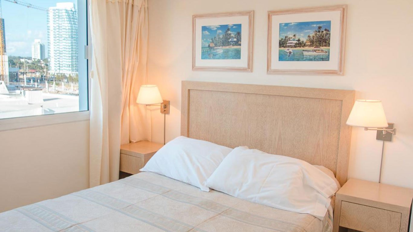Quarto de hotel no Hotel Sunset Beach em Punta del Este, com decoração simples e elegante, uma cama de casal com cabeceira bege, abajures de mesa de cabeceira e quadros de paisagens litorâneas na parede, complementados por uma vista da marina através da janela.