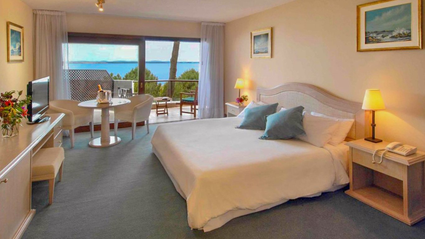 Quarto aconchegante no Hotel del Lago Golf & Art Resort em Punta del Este, com uma grande cama de casal, almofadas azul claro, decoração suave e uma mesa com bebidas de boas-vindas. Uma varanda ao fundo revela uma vista serena do lago.