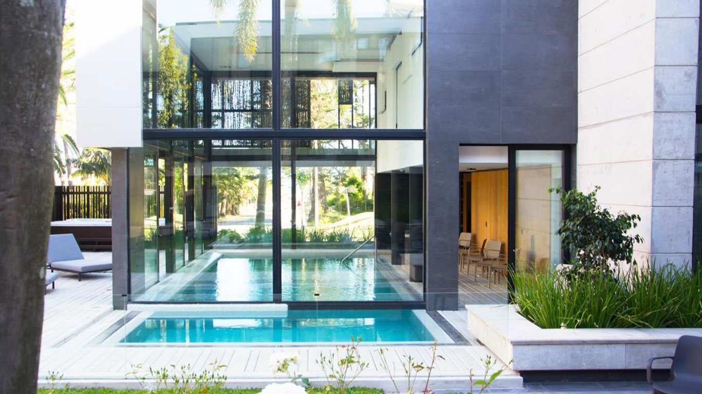 Design arquitetônico moderno do La Capilla em Punta del Este, com uma piscina de vidro transparente emoldurada por paredes de concreto e grandes janelas que refletem a vegetação circundante, oferecendo um oásis de tranquilidade e estilo.