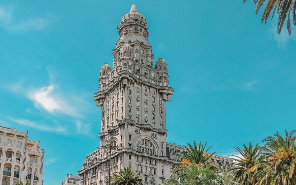 Vista do Palácio Savo durante o dia sob um céu azul claro. Este icônico edifício, localizado em Montevidéu, Uruguai, destaca-se por sua arquitetura neorrenascentista com detalhes ornamentais e uma torre proeminente. Palmeiras se elevam em primeiro plano, adicionando um toque tropical ao ambiente urbano.