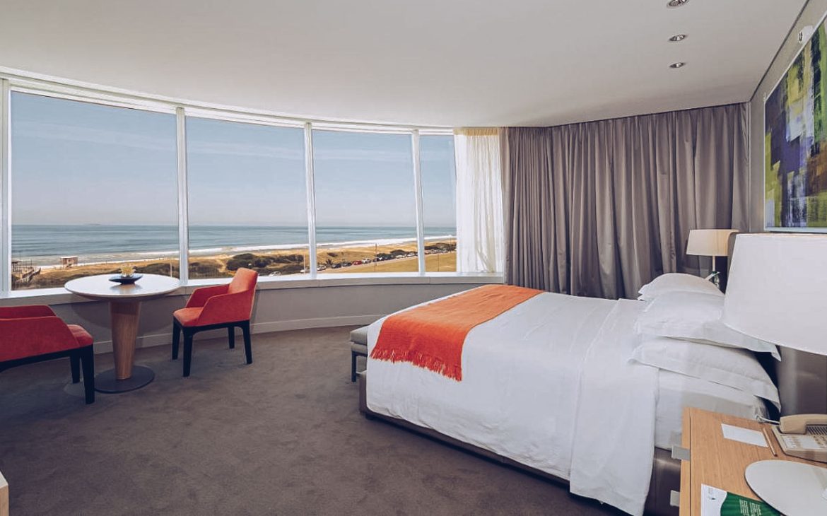 Quarto panorâmico do The Grand Hotel em Punta del Este, com paredes de vidro oferecendo uma vista ampla da praia e do horizonte. O interior é decorado com tons neutros, toques de cor laranja e mobília elegante para uma estadia confortável.