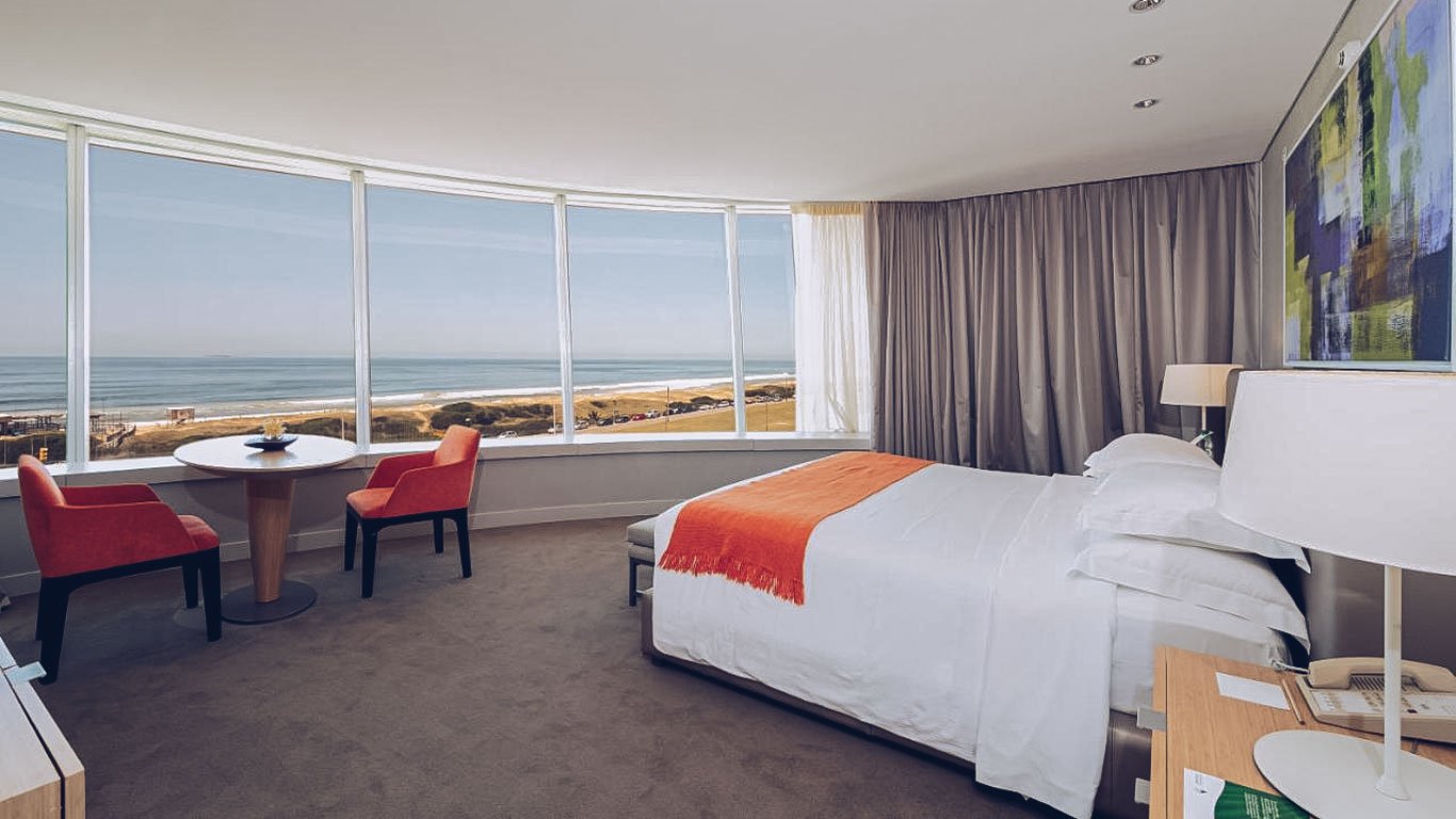 Quarto panorâmico do The Grand Hotel em Punta del Este, com paredes de vidro oferecendo uma vista ampla da praia e do horizonte. O interior é decorado com tons neutros, toques de cor laranja e mobília elegante para uma estadia confortável.
