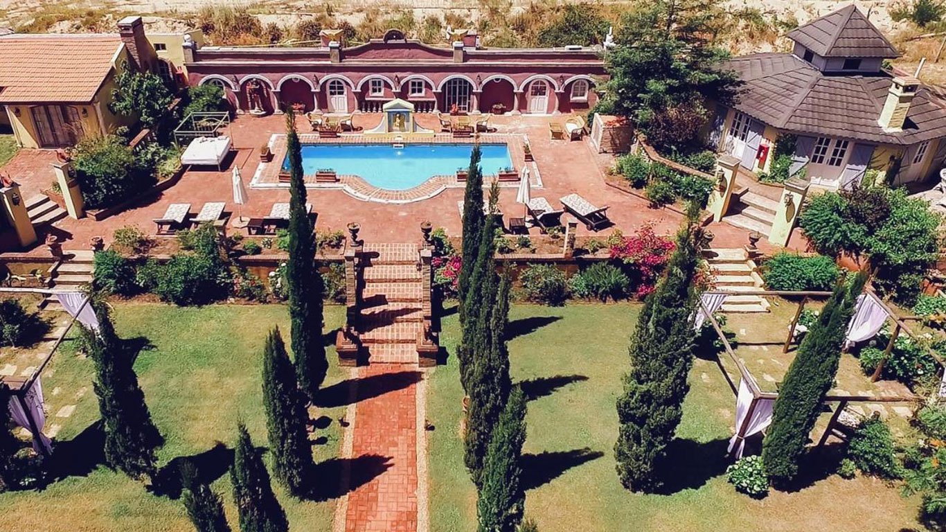 Vista aérea dos jardins simétricos e da piscina central do Villa Toscana Boutique Hotel, um dos melhores hotéis de Punta del Este, com um design reminiscente de uma villa italiana, cercado por ciprestes altos e áreas de lazer ao ar livre.
