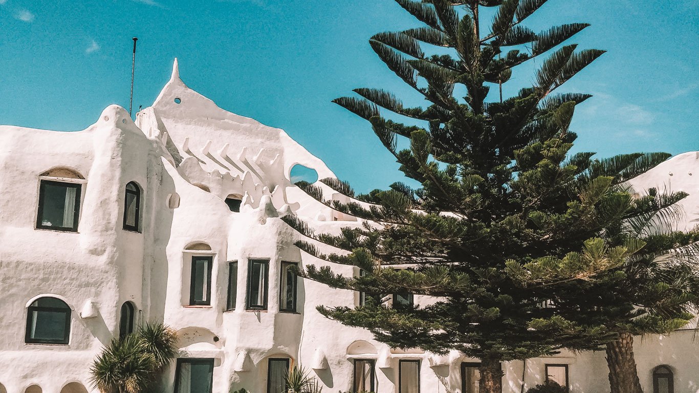 Vista da Casapueblo, um edifício branco com contornos curvos, sob um céu azul claro em Punta del Este. Um exuberante pinheiro se destaca em primeiro plano, contrastando com a textura lisa do edifício.