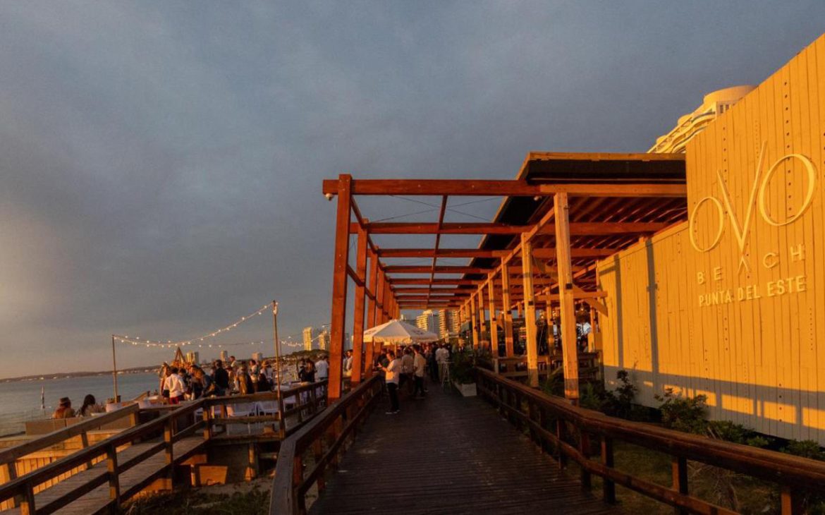 Entardecer no Enjoy Punta del Este com visitantes caminhando no calçadão de madeira decorado com luzes de corda, ao lado da parede exterior iluminada do local, que exibe seu logotipo.