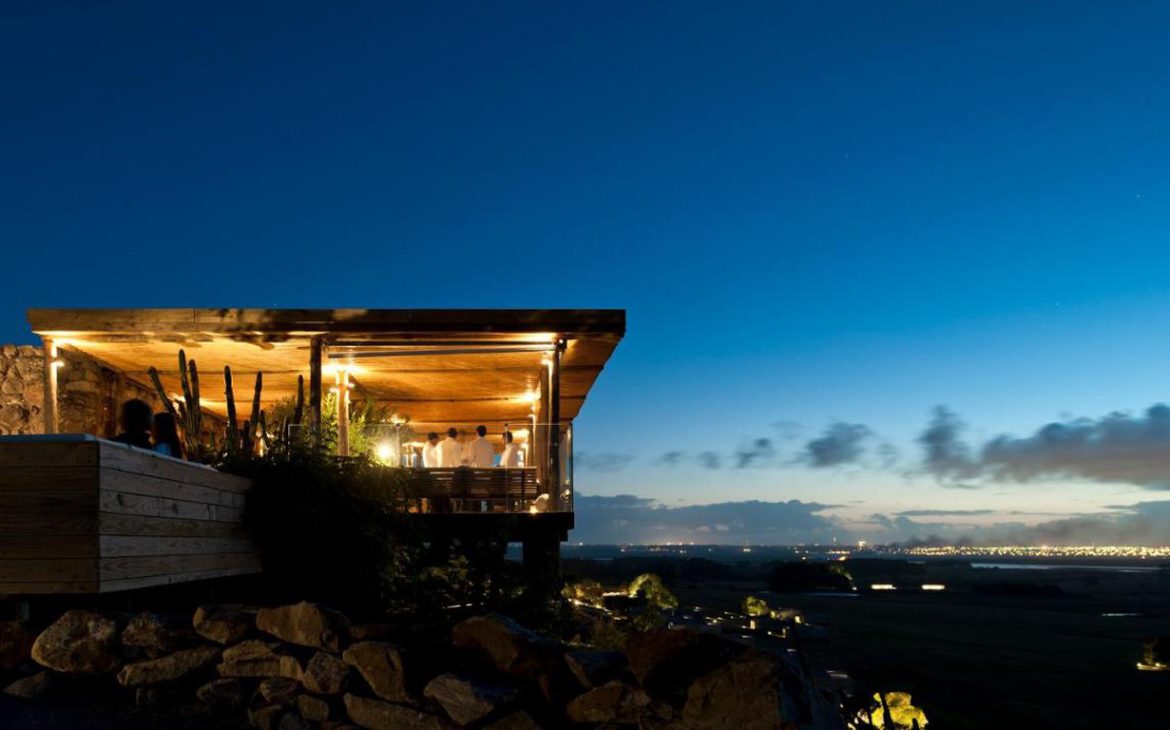 Vista noturna do Hotel Fasano Punta del Este, com destaque para o terraço iluminado, onde visitantes desfrutam da vista panorâmica da cidade iluminada ao fundo, sob um céu azul escuro.