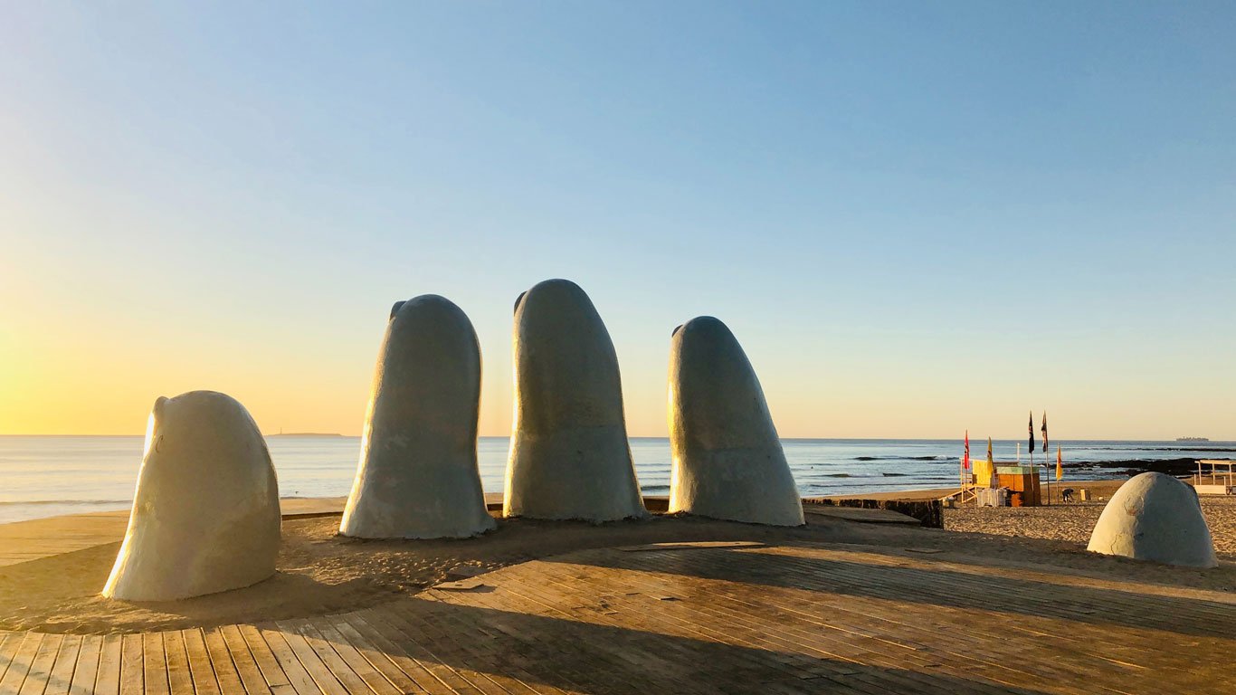 A icônica escultura 'La Mano' do artista chileno Mario Irarrázabal, representando cinco dedos gigantes emergindo da areia, na Praia Brava em Punta del Este, Uruguai, durante um tranquilo amanhecer com céu limpo.