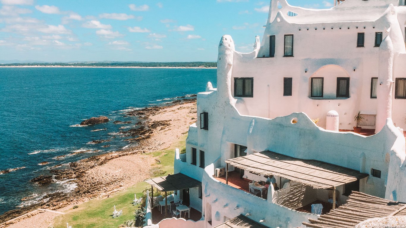 Casapueblo em Punta del Este com vista para um litoral rochoso, com as águas azuis do Oceano Atlântico ao fundo. As paredes caiadas e terraços do edifício oferecem um contraste marcante com a paisagem natural, convidando ao relaxamento e contemplação.