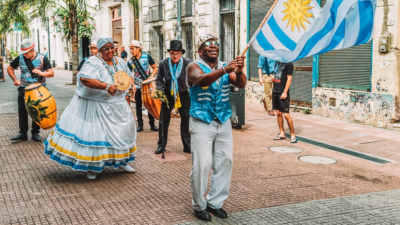 A imagem mostra um grupo animado de pessoas participando de um passeio cultural pelas ruas de Montevidéu, Uruguai. No centro, uma mulher vestida com um traje tradicional branco e azul dança ao som de tambores tocados por homens ao seu redor. Um homem entusiasmado, à frente do grupo, segura uma bandeira do Uruguai, transmitindo um ambiente festivo e vibrante.