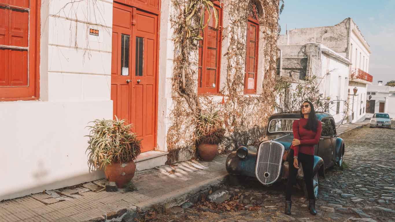 Mulher ao lado de um carro antigo em frente um imóvel em Colonia del Sacramento com portas alaranjadas e arbustos.