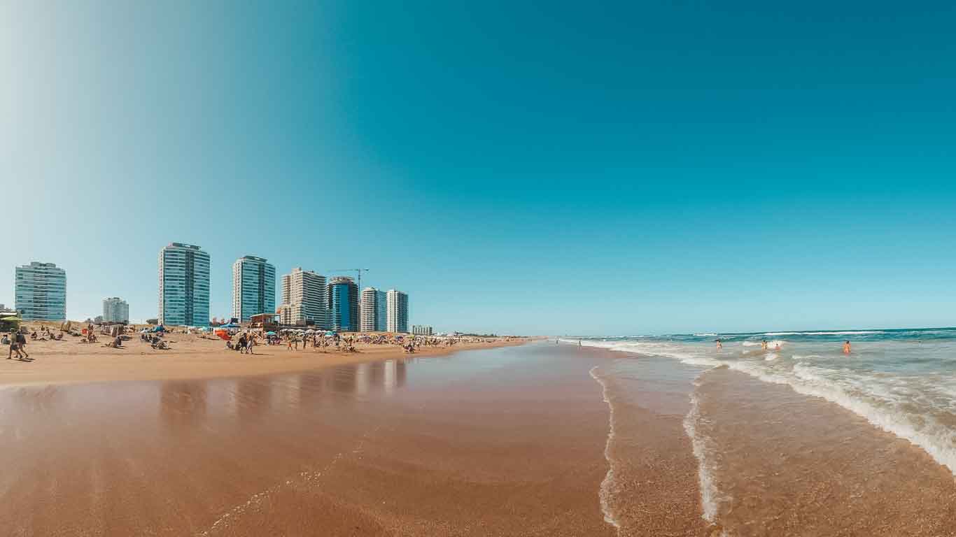 A movimentada Playa Brava em Punta del Este, Uruguai, exibe uma ampla praia de areia com visitantes aproveitando o sol e as ondas. Ao fundo, uma linha de modernos arranha-céus reflete o estilo de vida sofisticado da cidade costeira, sob um céu azul límpido.