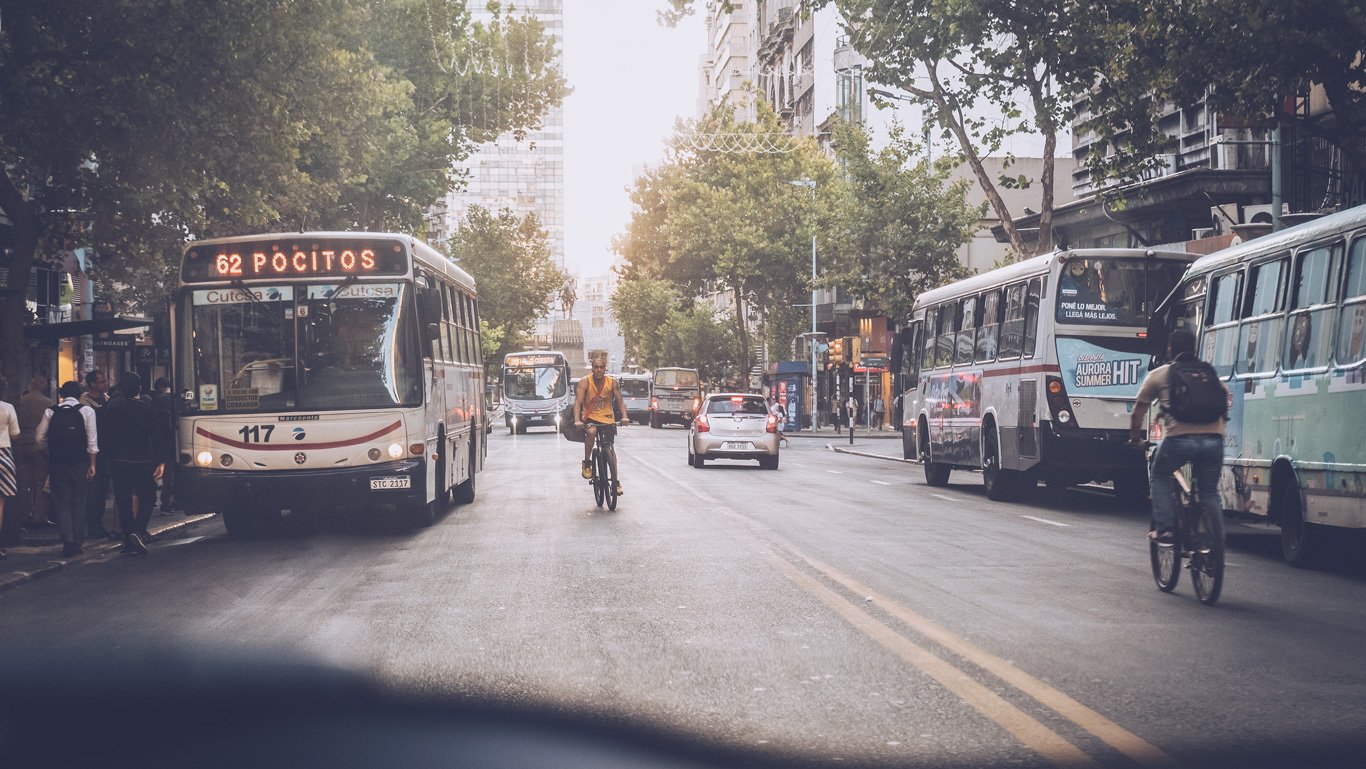 Cena de rua movimentada em Montevidéu, Uruguai, com um ciclista no centro e vários ônibus, incluindo um com a inscrição '62 POCITOS' e outro com um anúncio 'Aurora HIT Summer', destacando o cotidiano do deslocamento urbano.