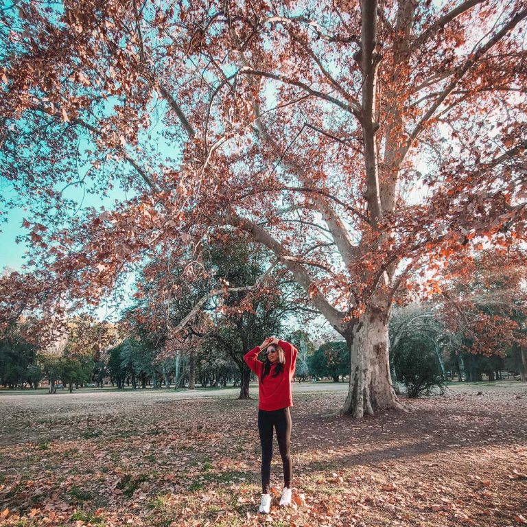 Mulher vestindo um casaco vermelho em pose descontraída no Parque General San Martín em Mendoza, durante o mês de junho, onde as folhas das árvores estão secas e o céu azul.