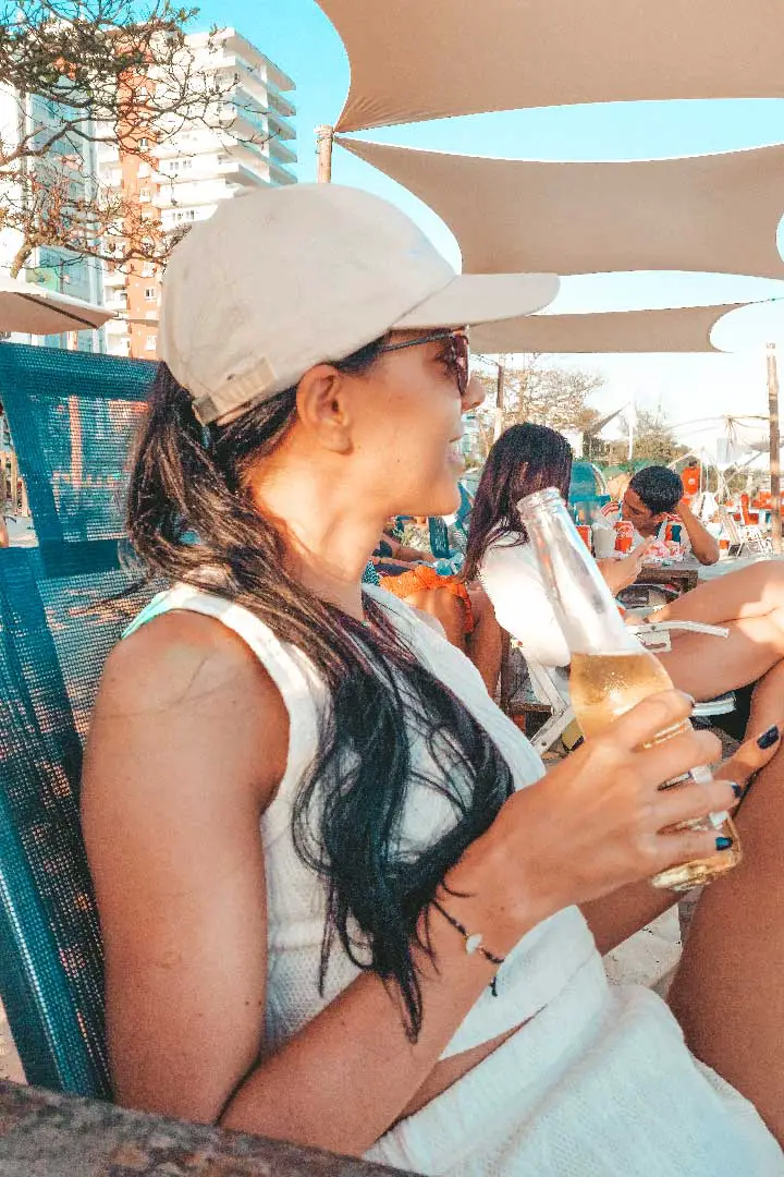 Mulher em um quiosque na praia da Barra da Tijuca, segurando uma bebida e sorrindo.