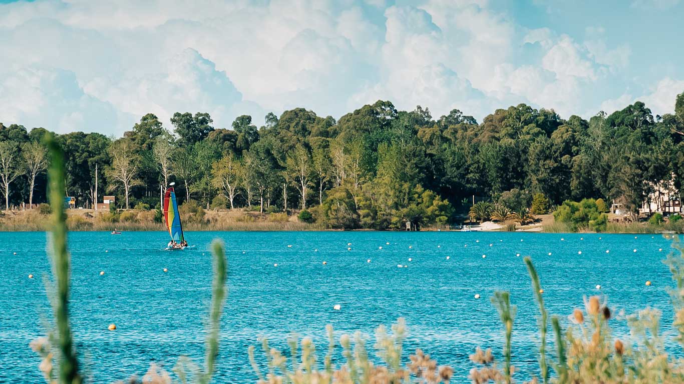 Vista panorâmica da Laguna del Sauce em Punta del Este, com águas azul-turquesa contrastando com a densa vegetação verde ao fundo. No centro da lagoa, uma pessoa pratica windsurf com uma vela colorida, adicionando um toque de cores vivas à cena tranquila.