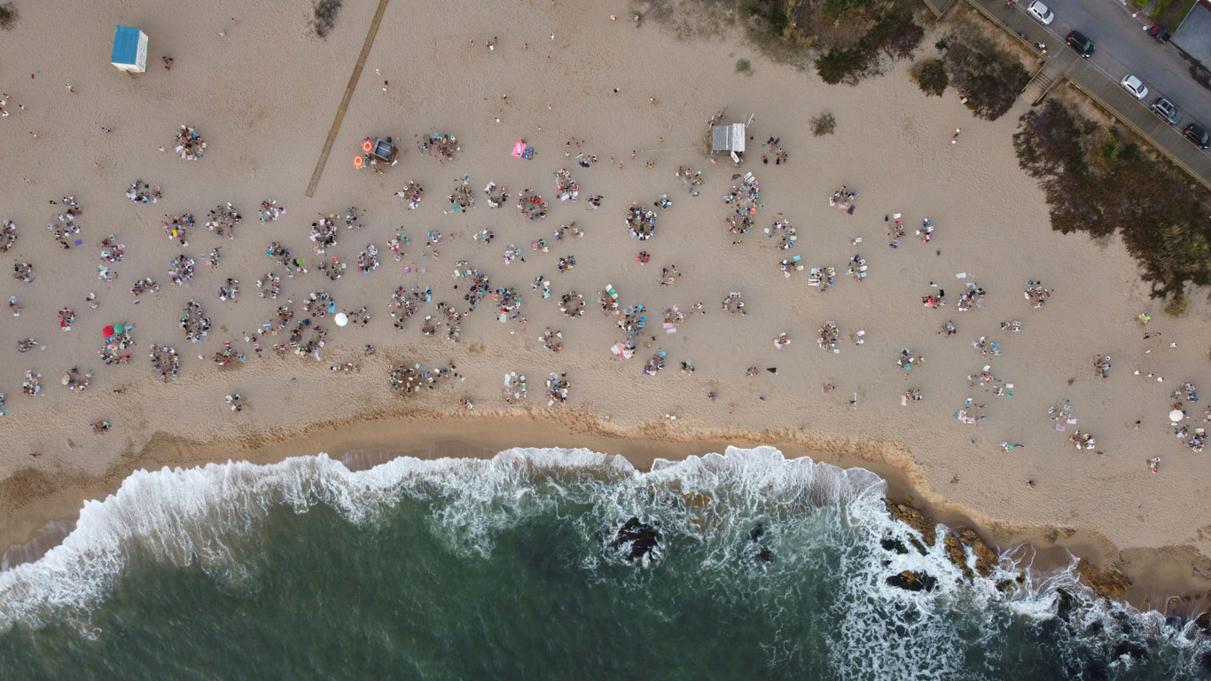 Vista aérea de uma cena movimentada de praia com pessoas reunidas na areia, guarda-sóis e toalhas de praia, ao lado de ondas quebrando na costa, com uma estrada e carros visíveis ao fundo.