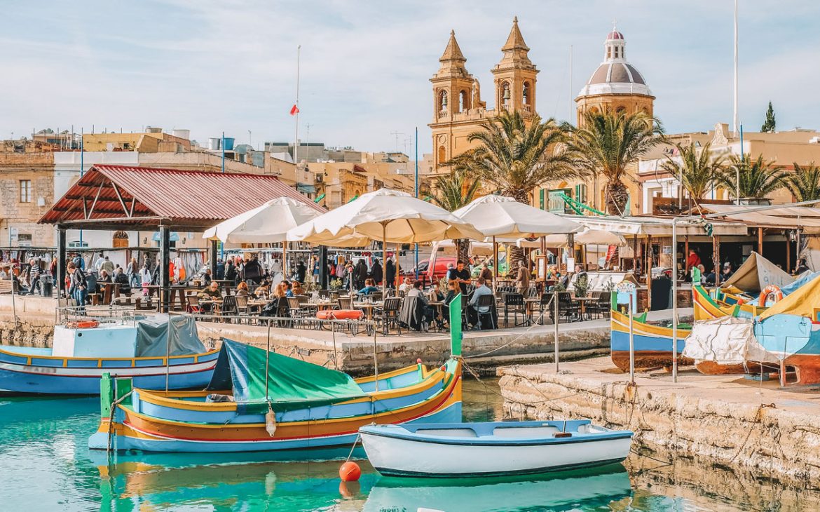 Cena movimentada à beira-mar em Marsaxlokk, em Malta, com os tradicionais barcos de pesca malteses coloridos, conhecidos como 'luzzus', flutuando nas águas azuis cristalinas. O fundo apresenta cafés ao ar livre movimentados com pessoas sob guarda-sóis brancos e arquitetura histórica, incluindo as icônicas torres da igreja.