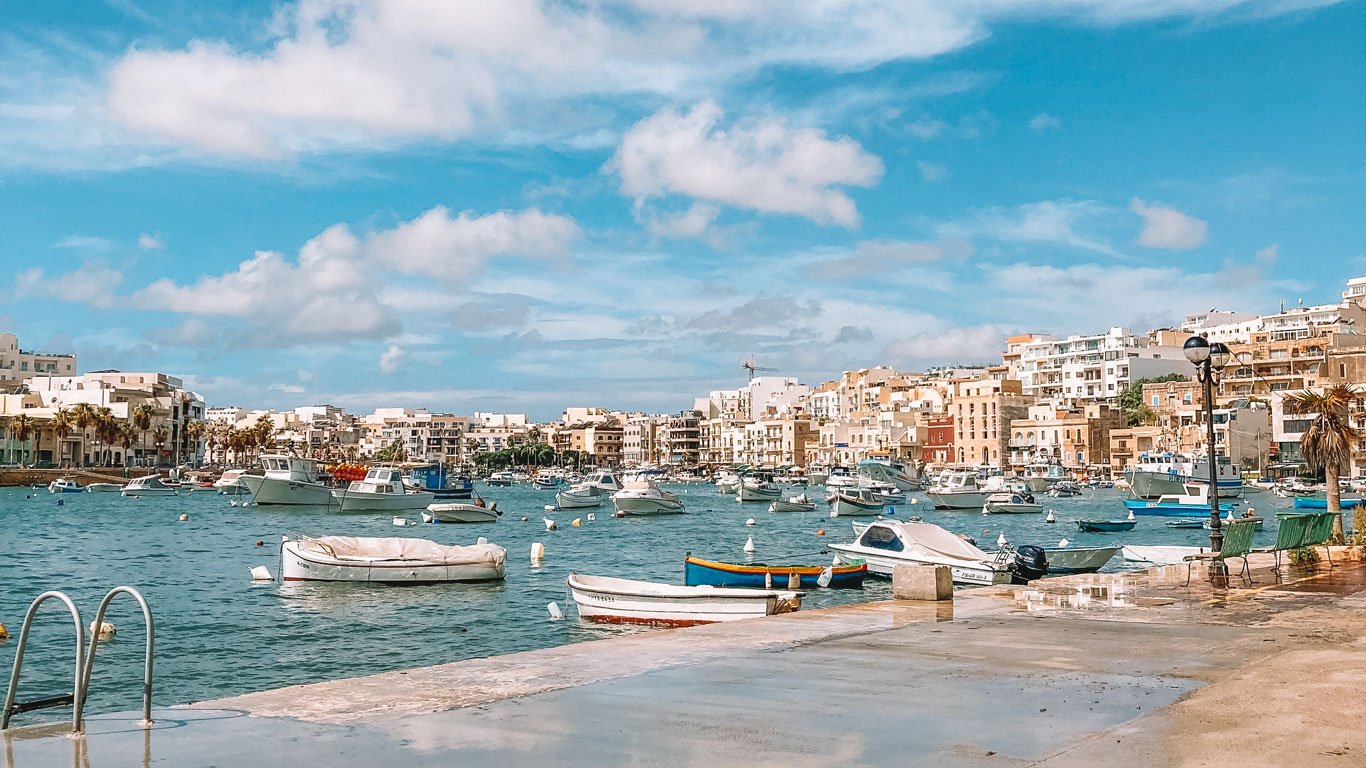 O mar azul de Marsaskala, em um dia de sol, com os barcos coloridos ancorados, ao fundo as casas e pequenos edifícios de tons claros compõem a paisagem.