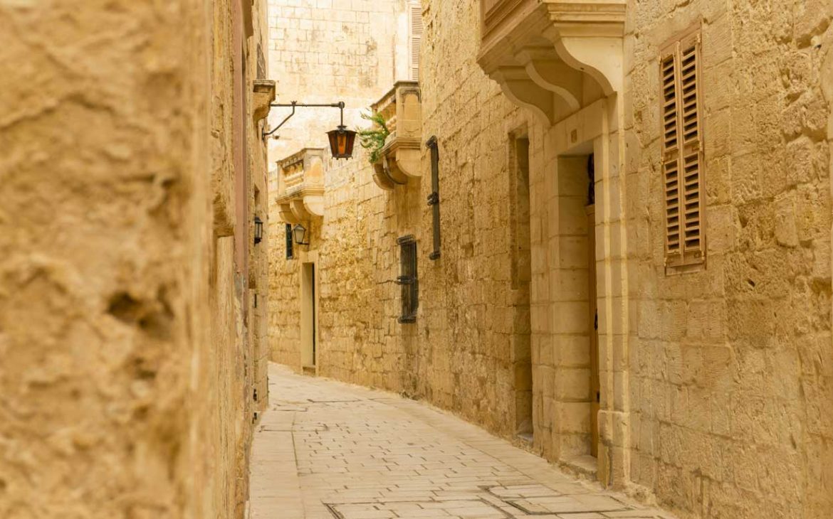 Uma rua estreita e tranquila na antiga cidade murada de Mdina, Malta, caracterizada pelas suas paredes de calcário, portas em arco e varandas tradicionais maltesas com persianas de madeira fechadas. O encanto intemporal da cidade é realçado pelos tons quentes da pedra e pelo verde ocasional dos vasos de plantas.