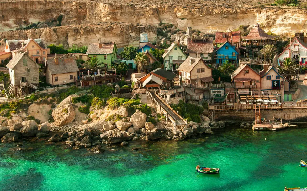 Popeye Village em Mellieha, a melhor opção de cidade de praia onde ficar em Malta. As águas incrivelmente verdes são emolduradas pela silhueta dos barcos de pescadores, enquanto ao fundo, as rochas acompanham o curso do mar, destacando-se pelas pequenas e coloridas casas de madeira que pontuam a paisagem rochosa.