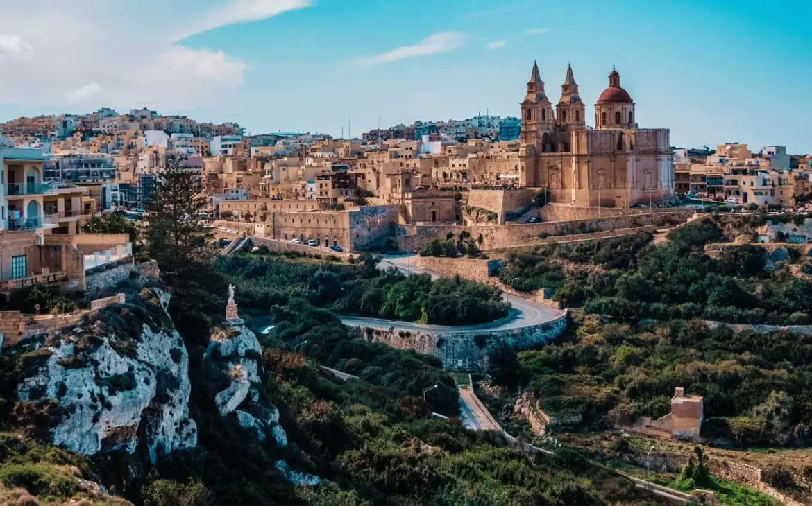 Vista panorâmica de Mellieha, um destino popular para ficar em Malta, apresentando a icónica Igreja Paroquial num cenário de casas geminadas e vales verdes, com estradas sinuosas que conduzem à serena cidade.
