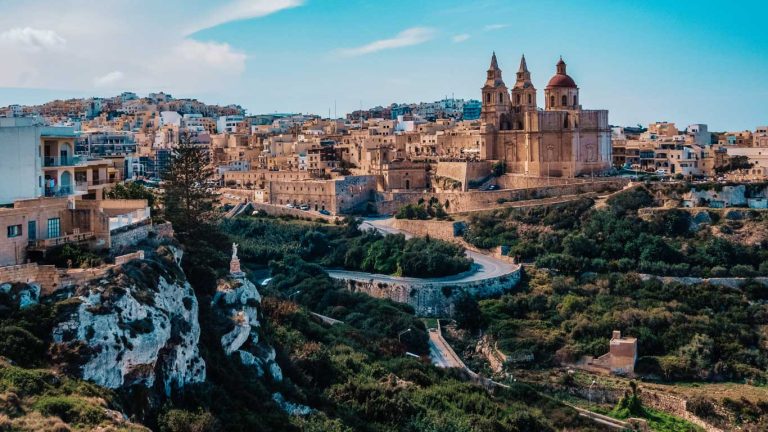 Vista panorâmica de Mellieha, um destino popular para ficar em Malta, apresentando a icónica Igreja Paroquial num cenário de casas geminadas e vales verdes, com estradas sinuosas que conduzem à serena cidade.