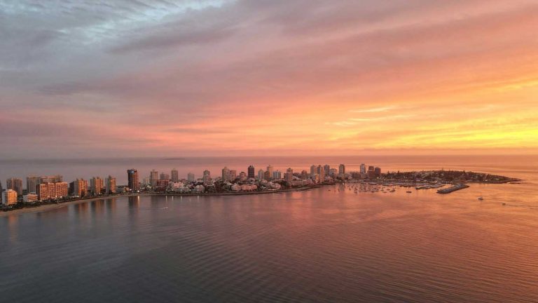 Vista aérea do pôr do sol tingindo o céu de tons rosados e alaranjados sobre Punta del Este, Uruguai. A paisagem urbana costeira é pontilhada com arranha-céus iluminados refletindo na calma água da baía, enquanto inúmeros barcos estão ancorados no porto.