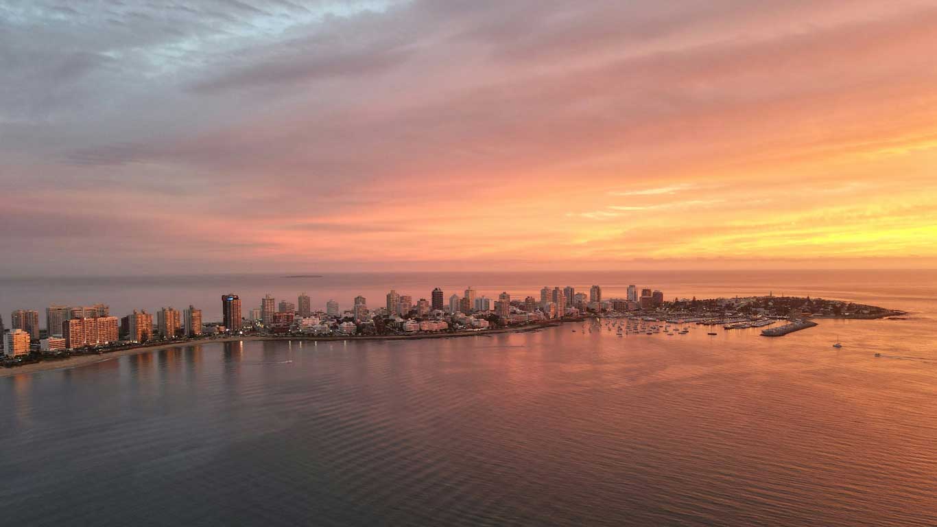 Vista aérea do pôr do sol tingindo o céu de tons rosados e alaranjados sobre Punta del Este, Uruguai. A paisagem urbana costeira é pontilhada com arranha-céus iluminados refletindo na calma água da baía, enquanto inúmeros barcos estão ancorados no porto.