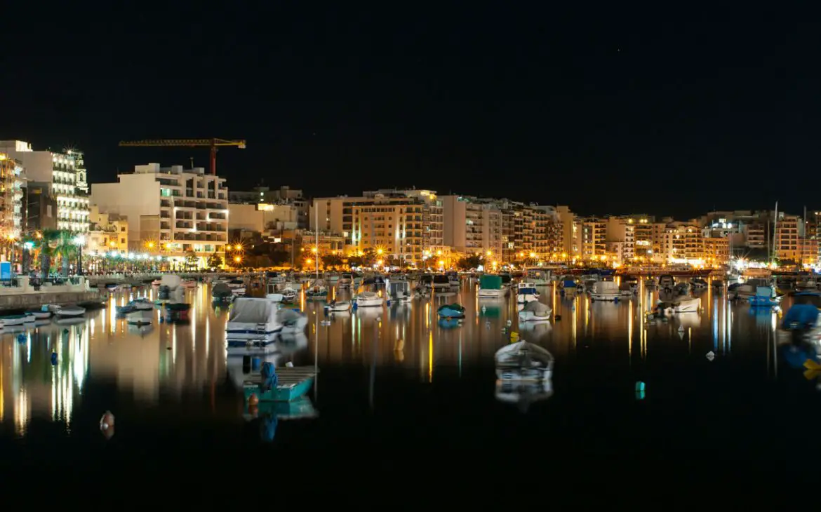 Baia de Sliema durante a noite, com as luzes da cidade refletindo no mar e iluminando os barcos ancorados, ao fundo os edifícios iluminados complementam a bela a paisagem.