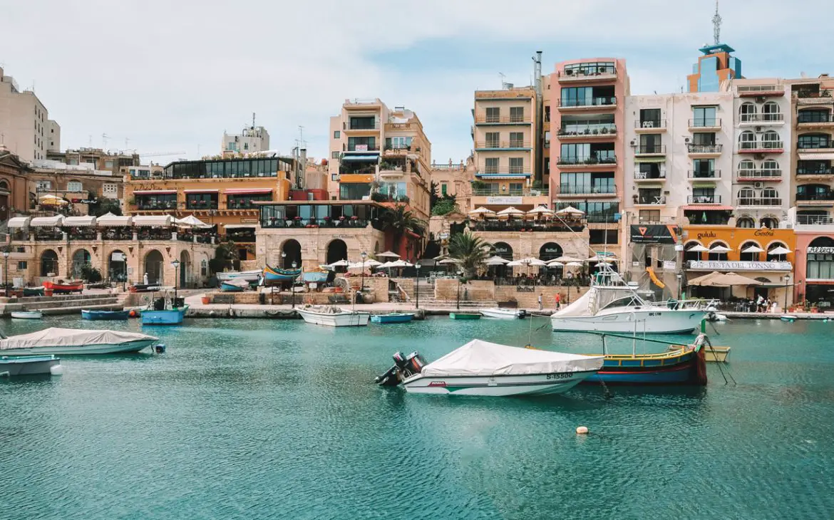 Um dia ensolarado na Baía de St. Julian's,o melhor lugar onde ficar em Malta, onde se destaca o azul intenso do mar salpicado de barcos coloridos, enquanto ao fundo, as pitorescas casas e edifícios oferecem um cenário vibrante e convidativo.