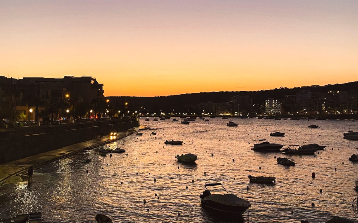 St Paul’s Bay ao anoitecer, com os últimos raios do sol iluminando o mar e os pequenos barcos que estão ancorados. Ao lado esquerdo as luzes começam a se acender na orla que acompanha o mar.