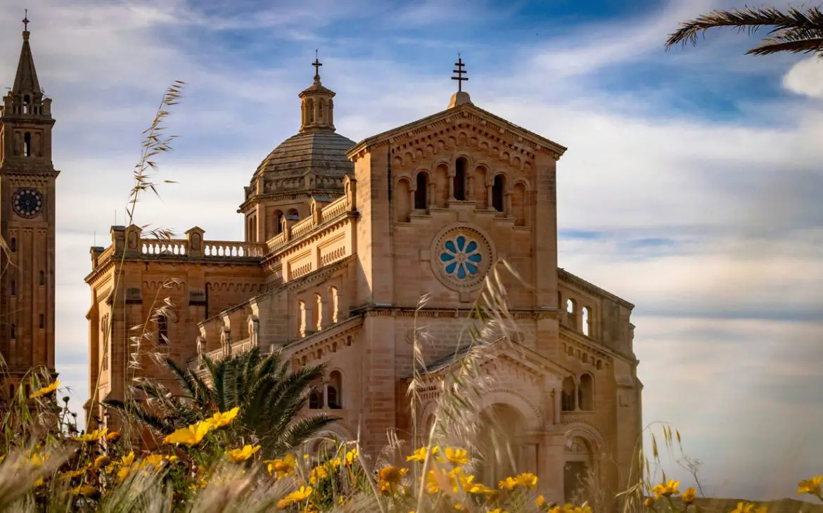 A Basílica de Ta' Pinu, em Gozo, ergue-se majestosamente em meio às flores silvestres, com sua arquitetura ornamentada e torres sineiras gêmeas se estendendo em direção a um céu azul suave, emanando uma sensação de paz e grandiosidade espiritual.