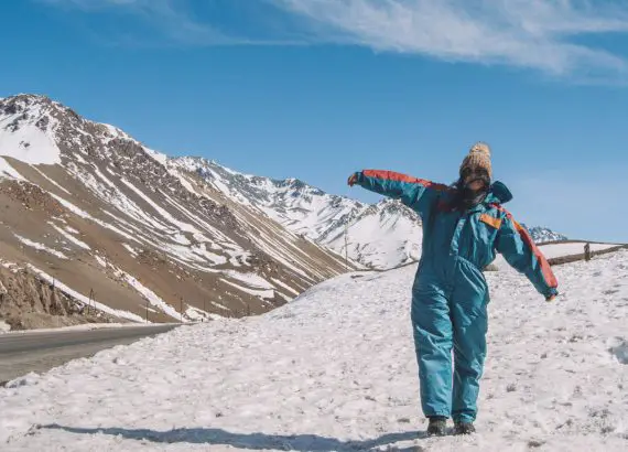 Turista alegre fazendo pose em uma paisagem nevada da Alta Montanha em Mendoza durante o mês de julho, vestindo roupas de inverno coloridas e um gorro, evidenciando a temporada de esqui.