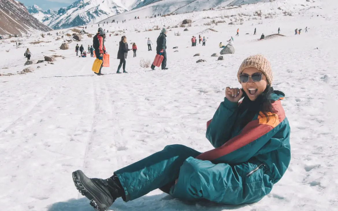 Turista sentada na neve sorrindo para a câmera em Mendoza, Argentina, com vestimenta apropriada para o frio, incluindo óculos escuros e gorro de lã, com outros visitantes e a paisagem montanhosa nevada ao fundo.