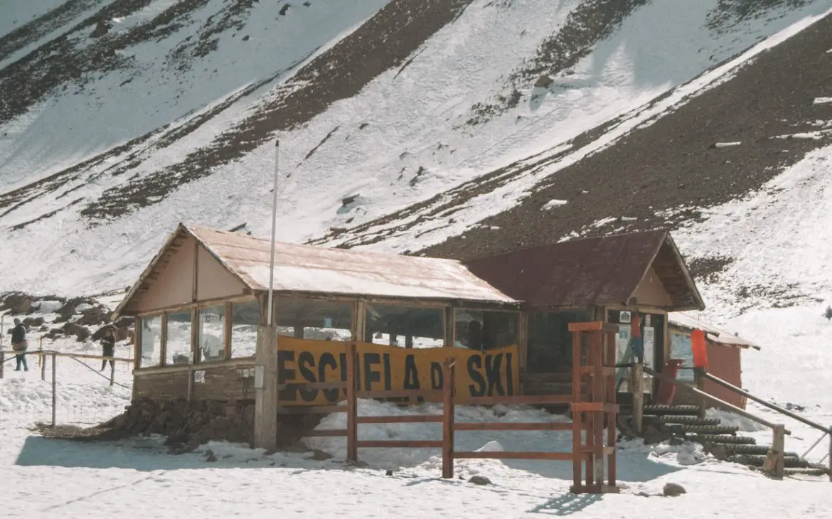 Escola de esqui em Mendoza com neve ao redor, sob céu azul claro, com as montanhas como pano de fundo e um sinal amarelo 'ESCUELA DE SKI' em destaque.