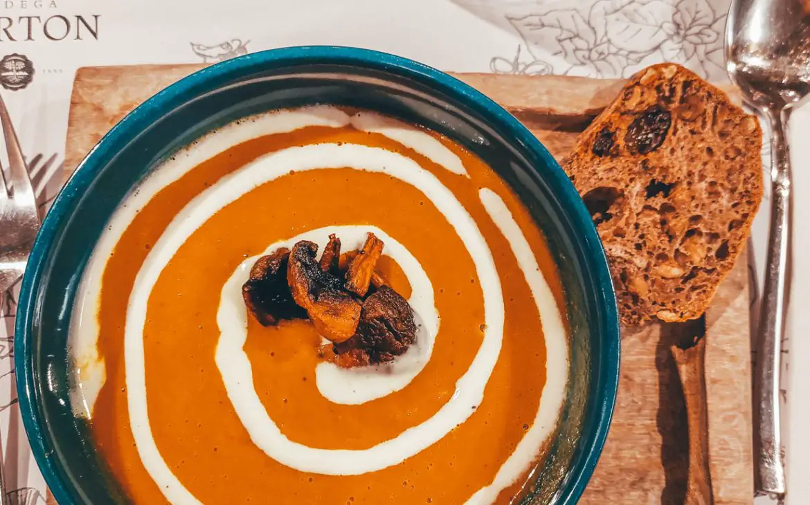 Prato de sopa cremosa de cenoura servida em um prato rústico com detalhes azuis na borda, acompanhada por uma fatia de pão de nozes e passas sobre em uma tábua de madeira, num restaurante de Mendoza.