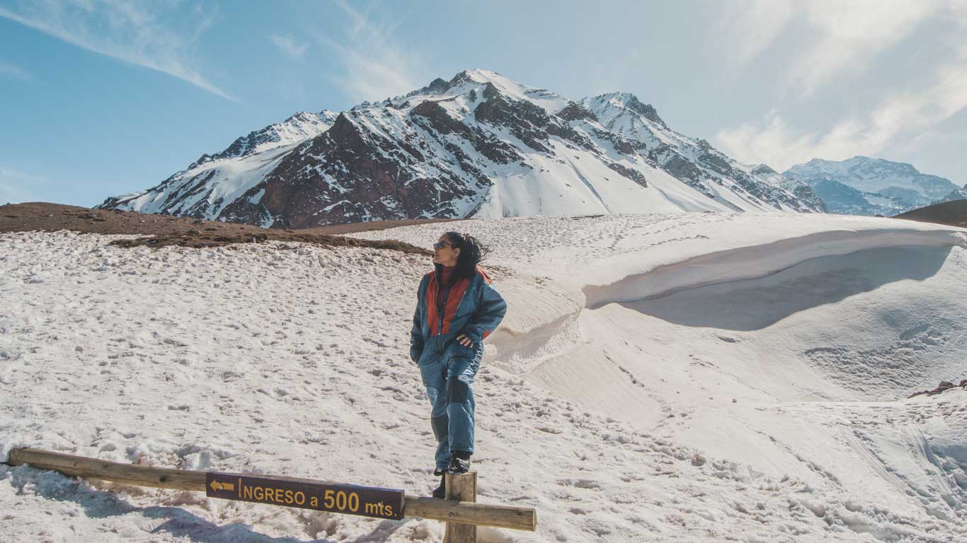Uma mulher de macacão de inverno azul e vermelho está em pé sobre uma cerca de madeira com uma placa que diz "INGRESO a 500 mts." ao fundo, uma montanha nevada em Mendoza, Argentina, sob um céu azul claro.